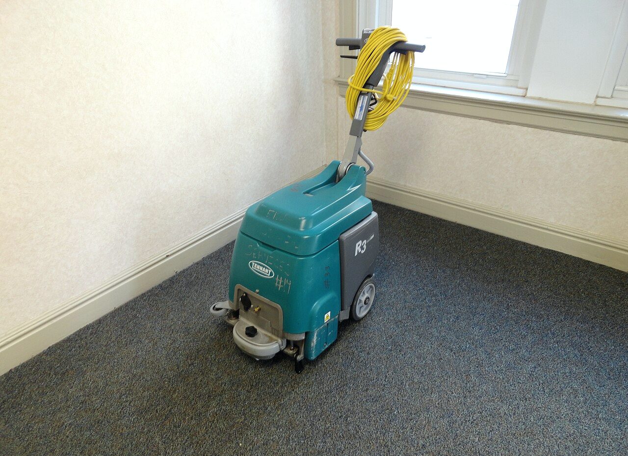 PLACEHOLDER: a wheeled carpet extractor with its cord coiled on the handle, standing on the carpet of an emptied room.