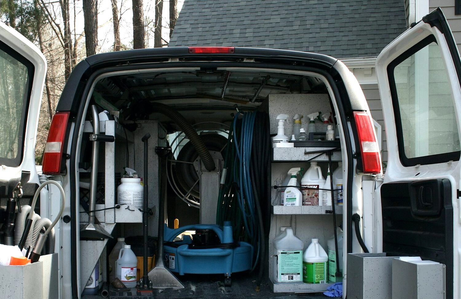 PLACEHOLDER: a carpet cleaning van with its rear doors open, showing coiled vacuum and solution hose, an air mover and shelves of chemicals.